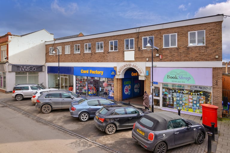 Street view of a row of brick storefronts with parked cars, including a candy factory shop and bookstore with a red postbox