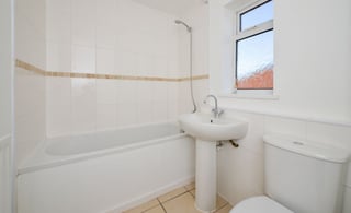 Clean, modern bathroom with white tile, bathtub with glass shower enclosure, pedestal sink, toilet, and small window with autumn view