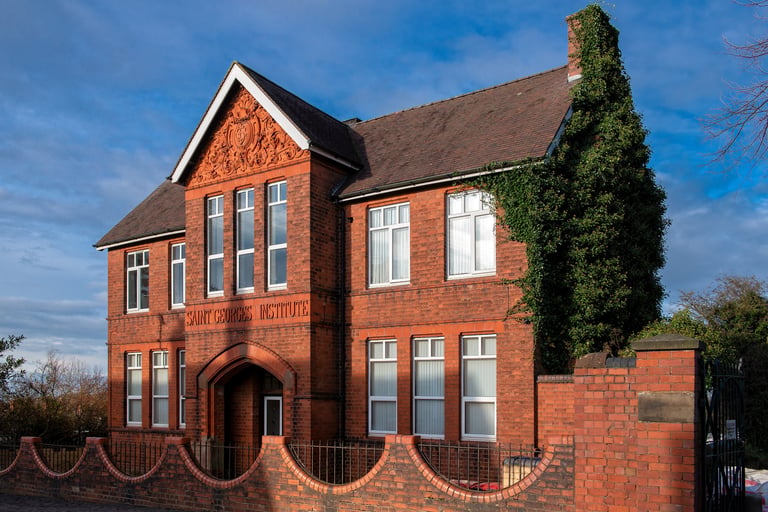 Red brick Victorian building with ivy-covered tower, arched entrance, and decorative fence against blue sky