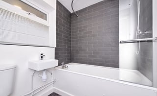 Modern bathroom with white toilet and sink, dark gray subway tile walls, white bathtub with glass shower partition, and small window above sink