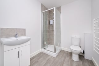 Modern bathroom with white curved sink vanity, glass shower enclosure, toilet, heated towel rack, and gray wood-look flooring