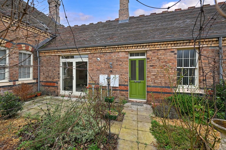 Victorian brick cottage exterior with bright green door, multiple windows, pale stone courtyard, and overgrown garden plants