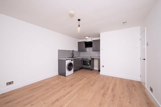 Modern apartment kitchen and living area with white walls, wooden laminate flooring, kitchenette with washing machine, and black appliances